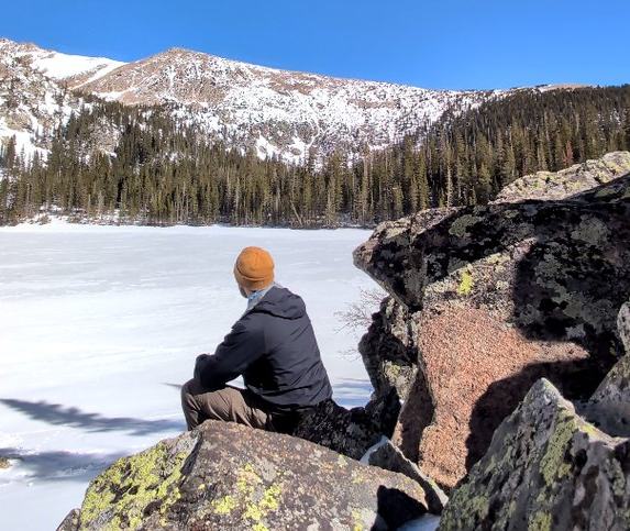 A person in an orange beanie sitting on lakeside boulders, looking out over a frozen alpine lake toward snowy peaks